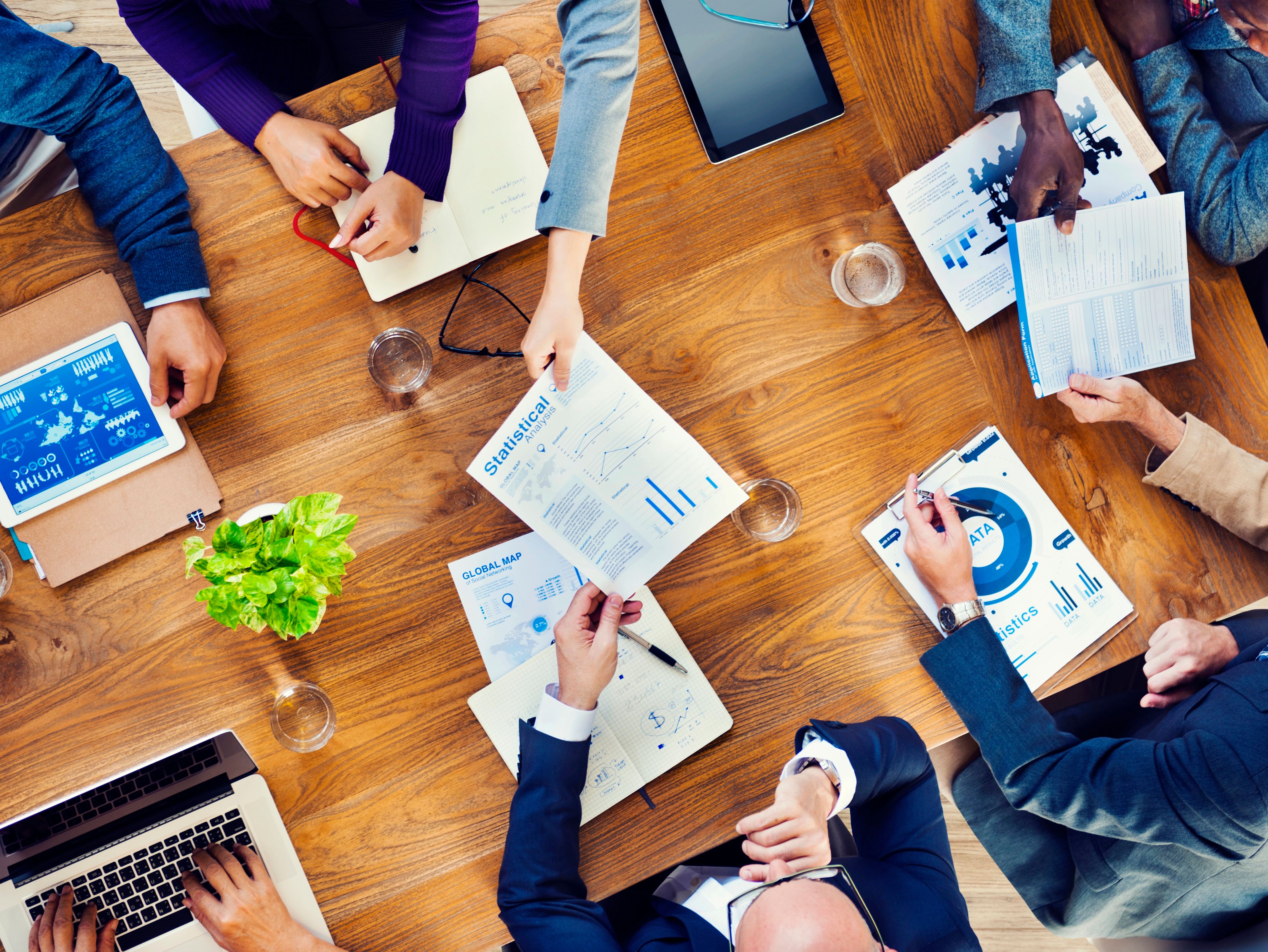 Ein Tisch von oben, an dem mehrere Menschen sitzen, die sich Dokumente zeigen. Auch digitale Geräte liegen auf dem Tisch.
