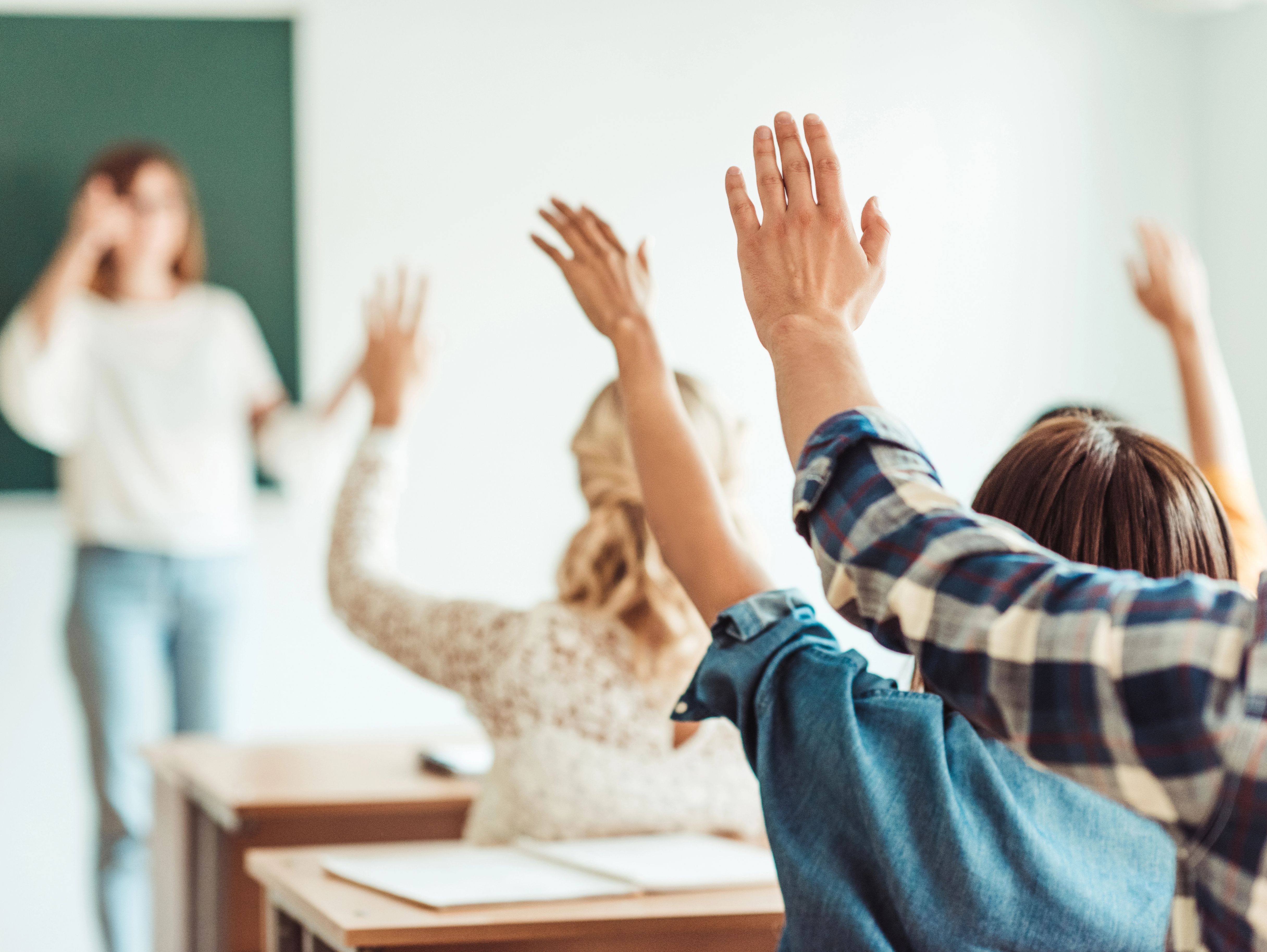 Auf dem Bild sind Schülerinnen und Schüler zu sehen, die ihre Hände in die Höhe halten. Verschwommen sieht man einen Tafel und eine weibliche Person, vermutlich die Lehrerin