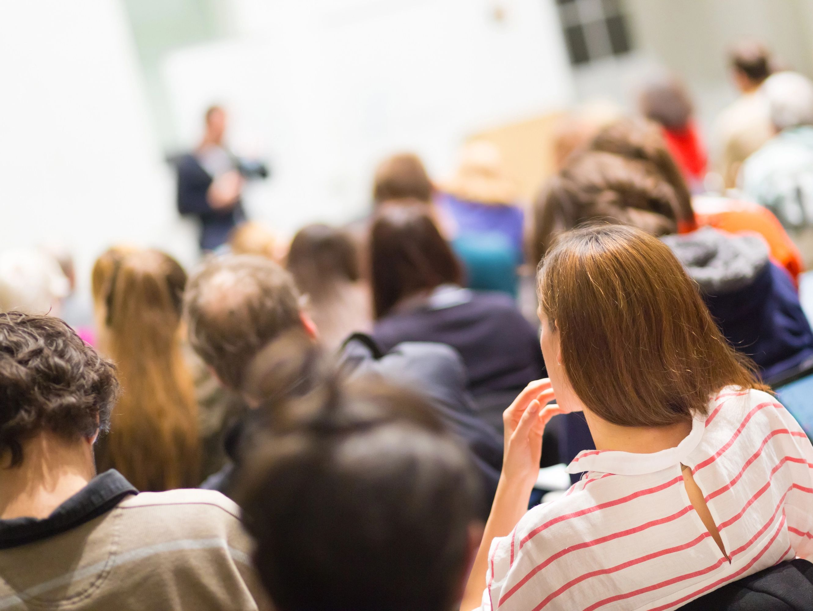 Im Hintergrund steht ein einzelner Mensch, der verschwommen zu sehen ist. Seine Zuhörerinnen und Zuhörer sieht man von hinten.